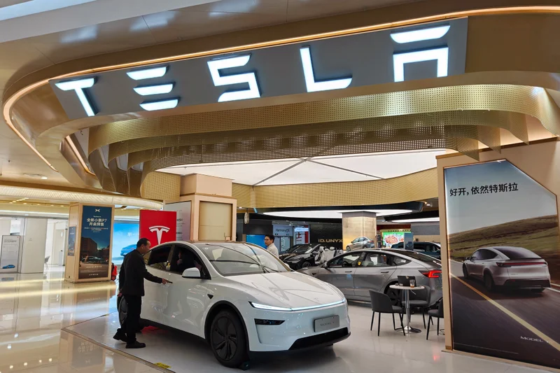 A white Tesla vehicle displayed in a modern showroom, with a man inspecting it under prominent Tesla signage.