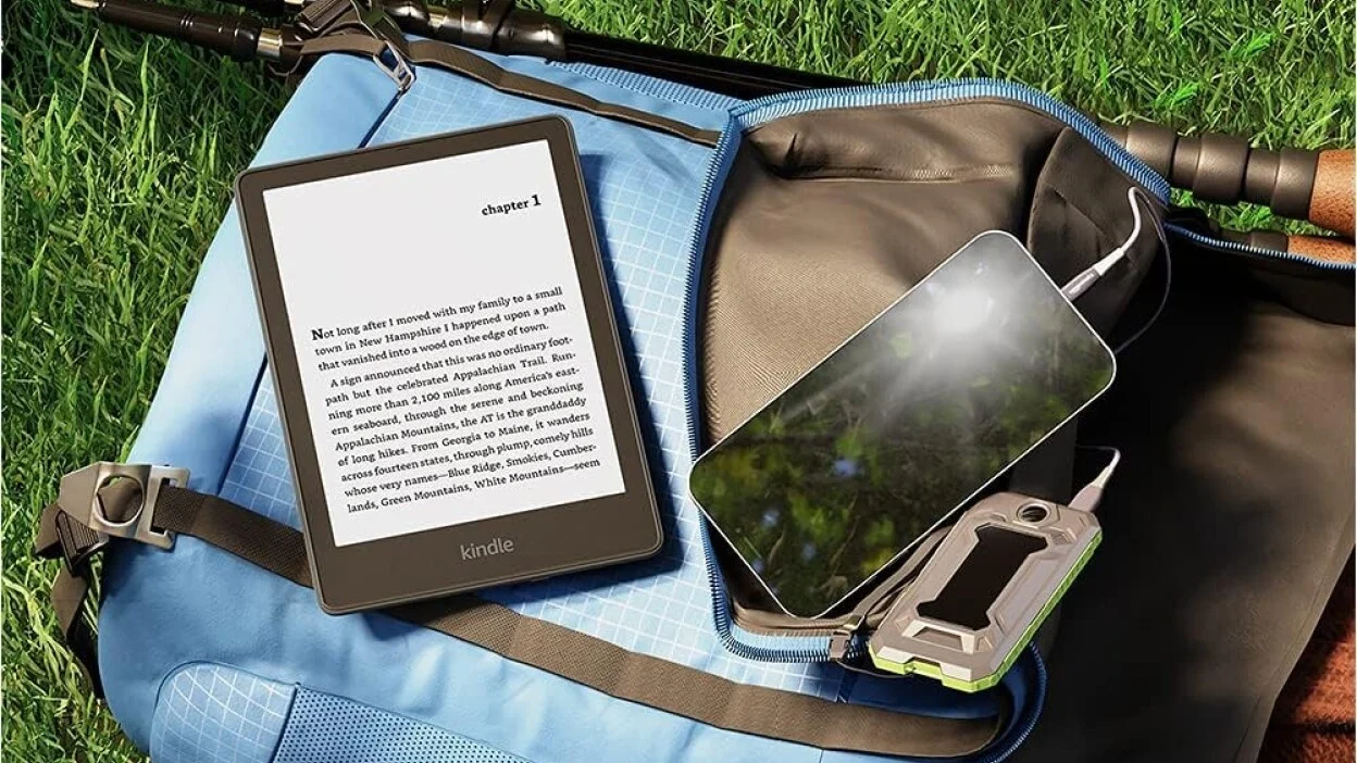 a kindle paperwhite sits on a backpack in the grass with a cell phone next to it