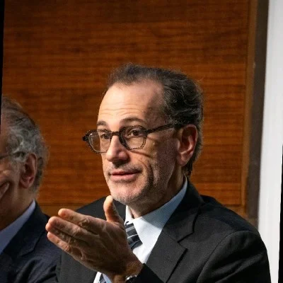 A middle-aged man in glasses gestures while speaking, wearing a suit and tie, with a wooden background visible.