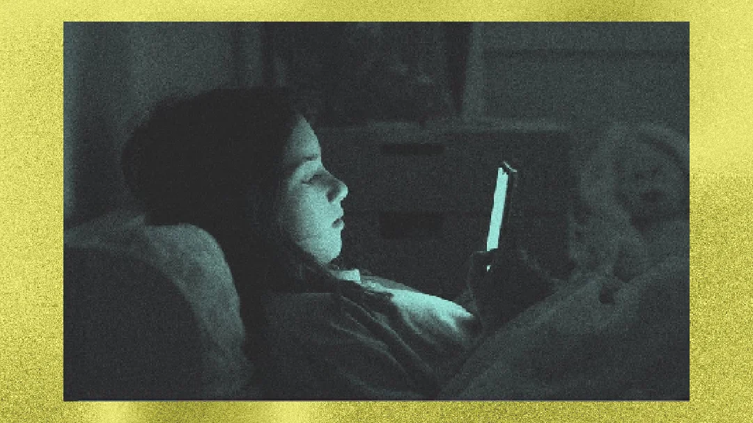 A person in bed looking intently at a phone screen, illuminated by its glow in a dimly lit room.