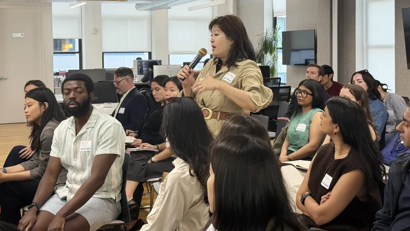 A woman in a beige outfit speaks into a microphone while seated audience members listen attentively in a modern office space.