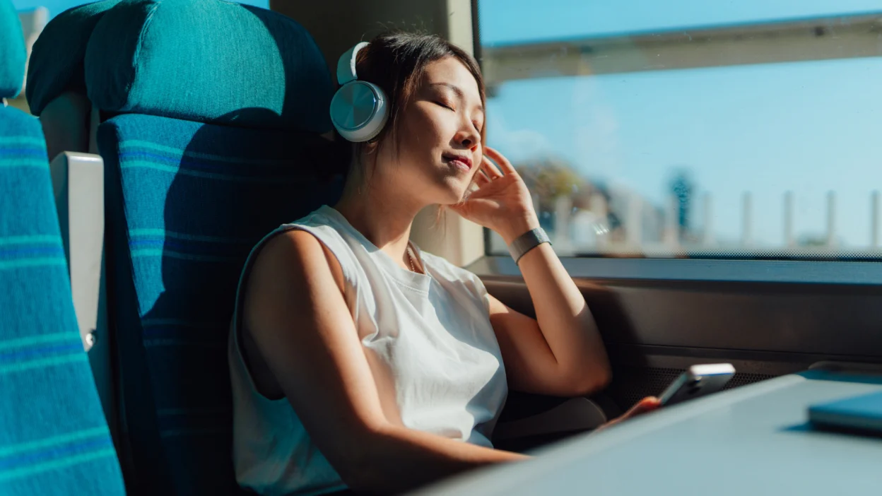 Young Asian woman with eyes closed, listening to music with wireless headphones in train.