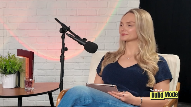 A woman with long blonde hair sits in a cream-colored chair during a podcast-style interview, holding a tablet and facing a microphone on a boom stand. She wears a navy blue top and jeans. Beside her is a small round table with a potted plant, several books, and a glass of pink sparkling drink. The background features a white brick wall with soft colored lighting and a black curtain, with a TechCrunch BuildMode logo in the bottom-right corner.