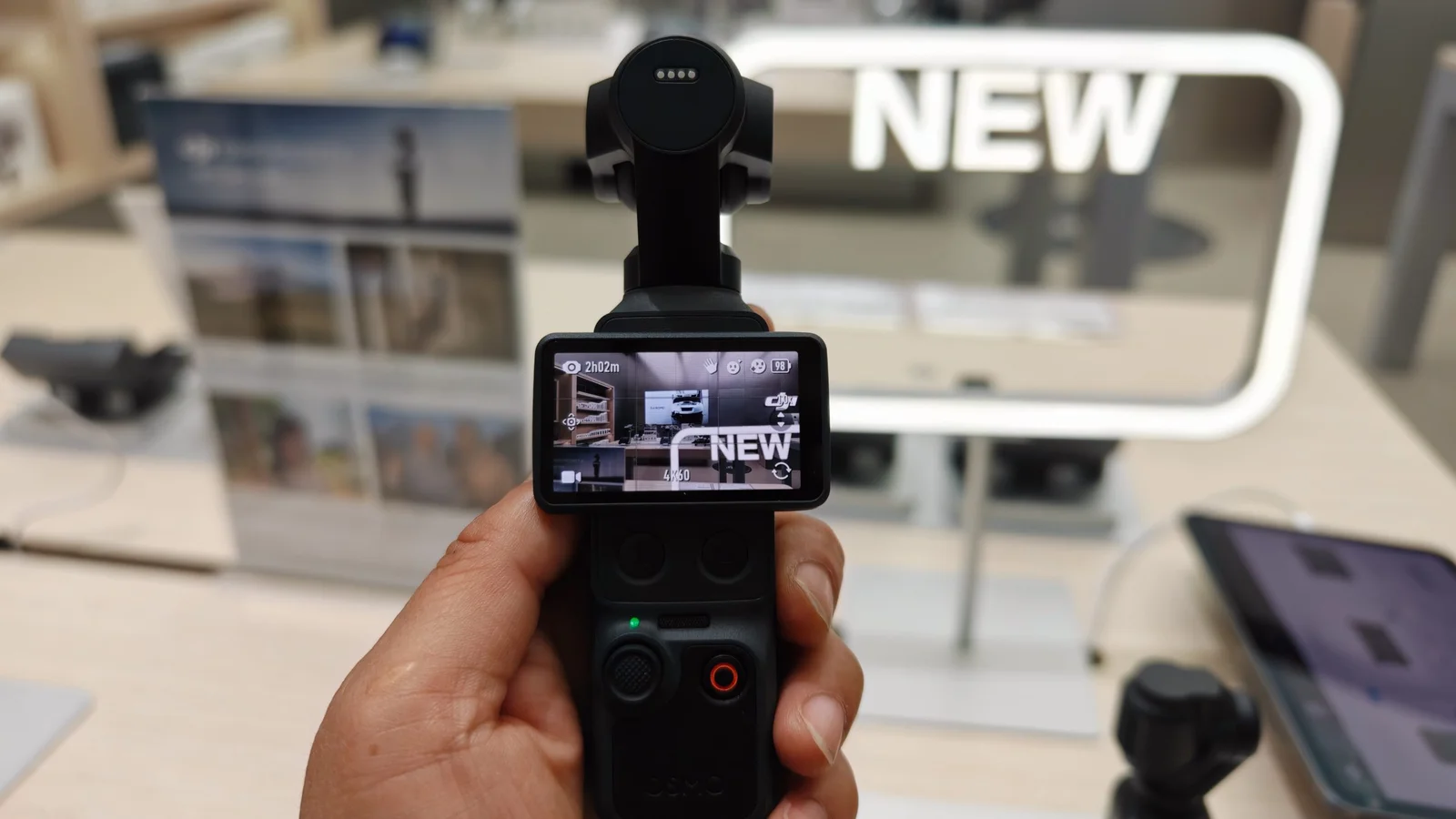 A person holds a handheld camera with a display showing the word "NEW," in a store with promotional materials in the background