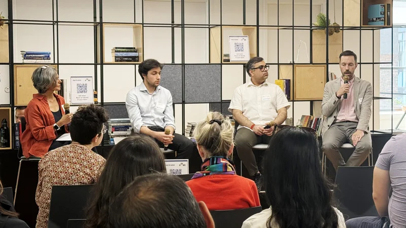 A panel discussion featuring four speakers seated in front of a bookshelf, with an audience visible in the foreground.