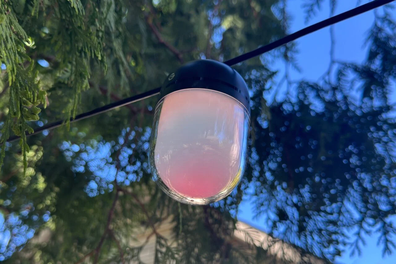 A Govee Outdoor Chromatic String Light bulb displaying a pink and blue gradient, during the day.