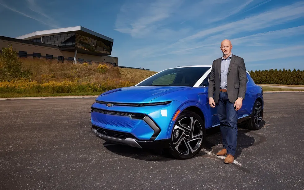 Photo of a bald man in a sports coat and jeans standing next to a royal blue car.