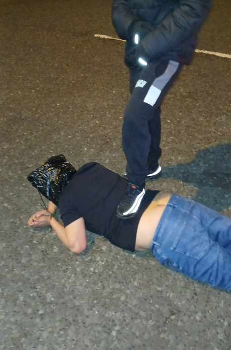 Many lying on the floor with a bag over his head and another man with his foot on his back