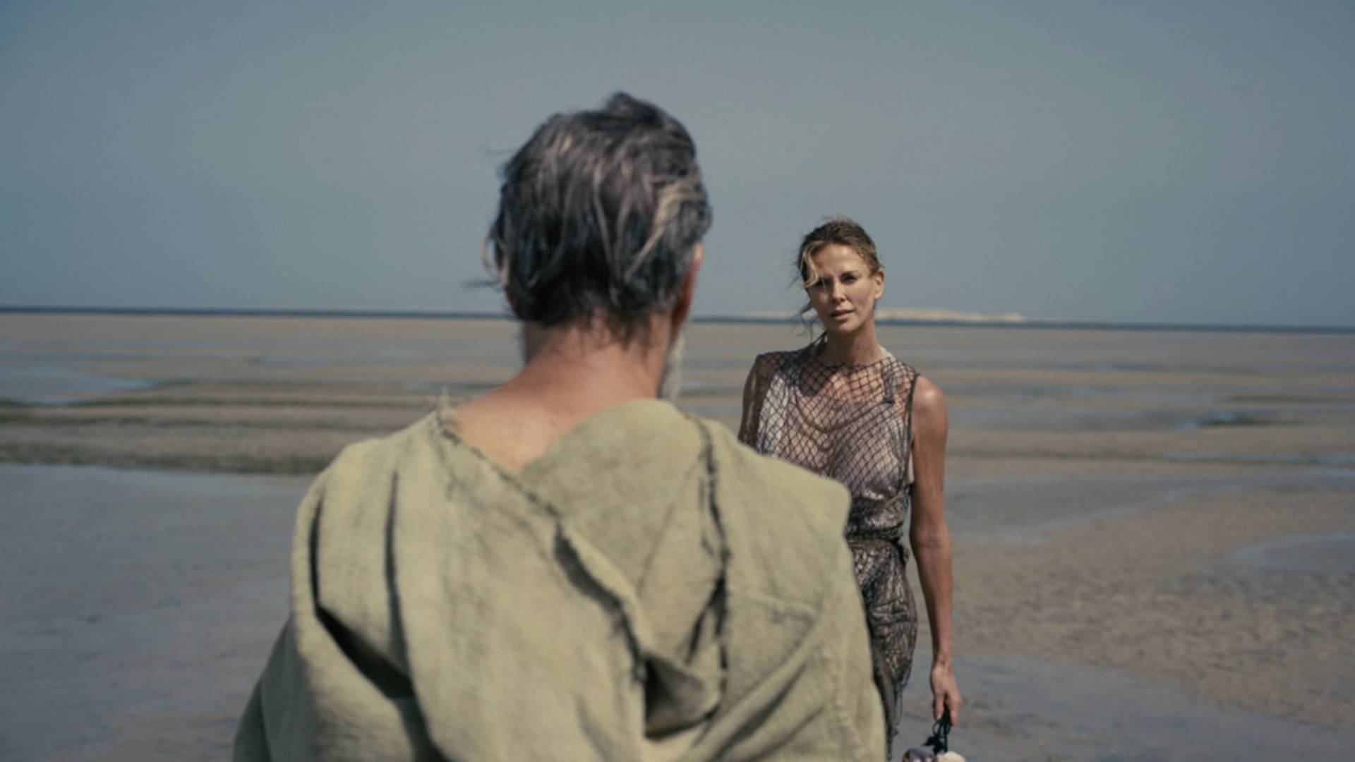 Charlize Theron as Calypso in The Odyssey, standing on a beach with Matt Damon's Odysseus